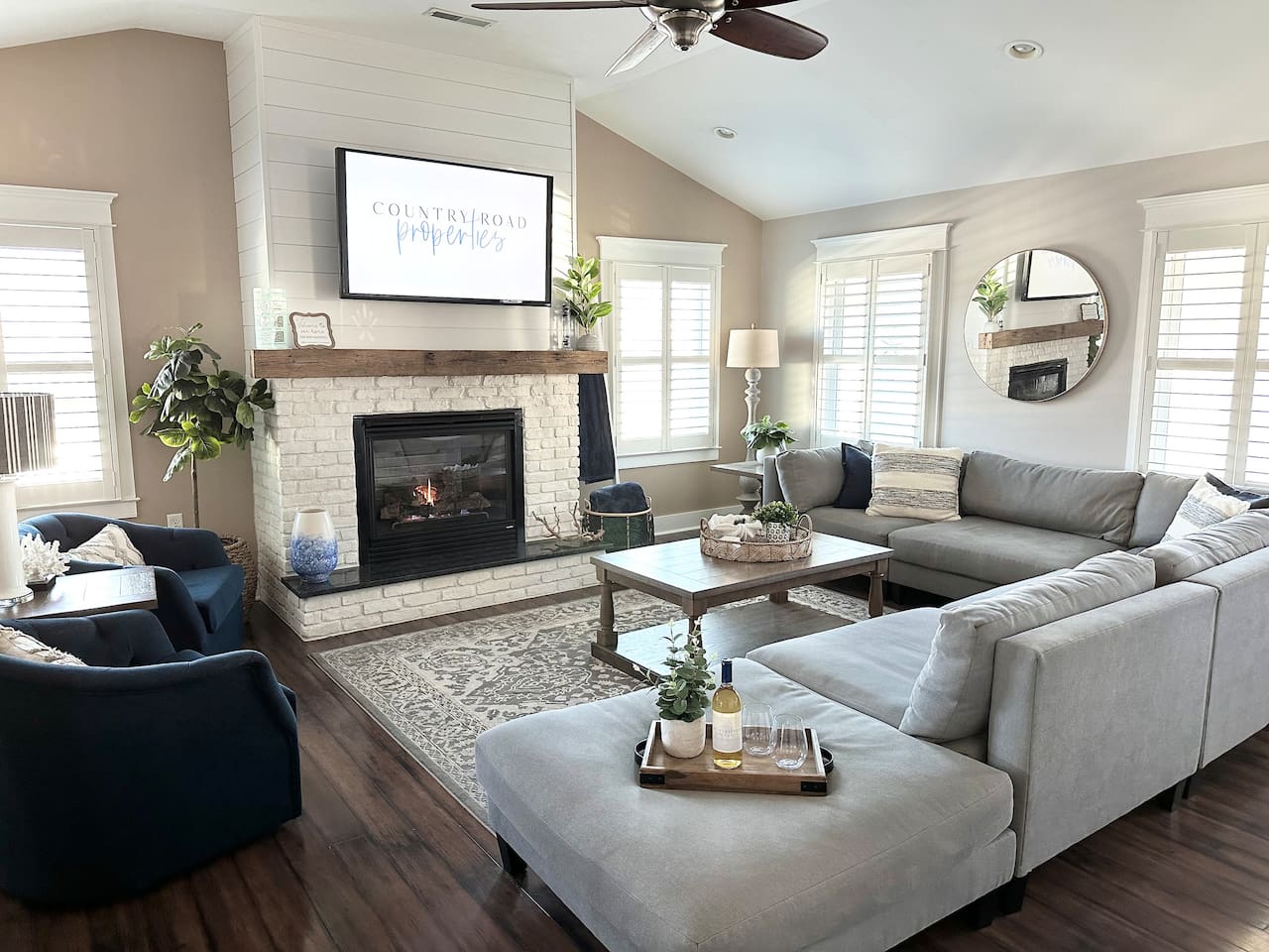 Gather in this sun-drenched living room, featuring vaulted ceilings and a cozy white brick fireplace. The sprawling grey sectional and navy accent chairs offer plenty of space to relax and watch a movie after a long day at the beach.