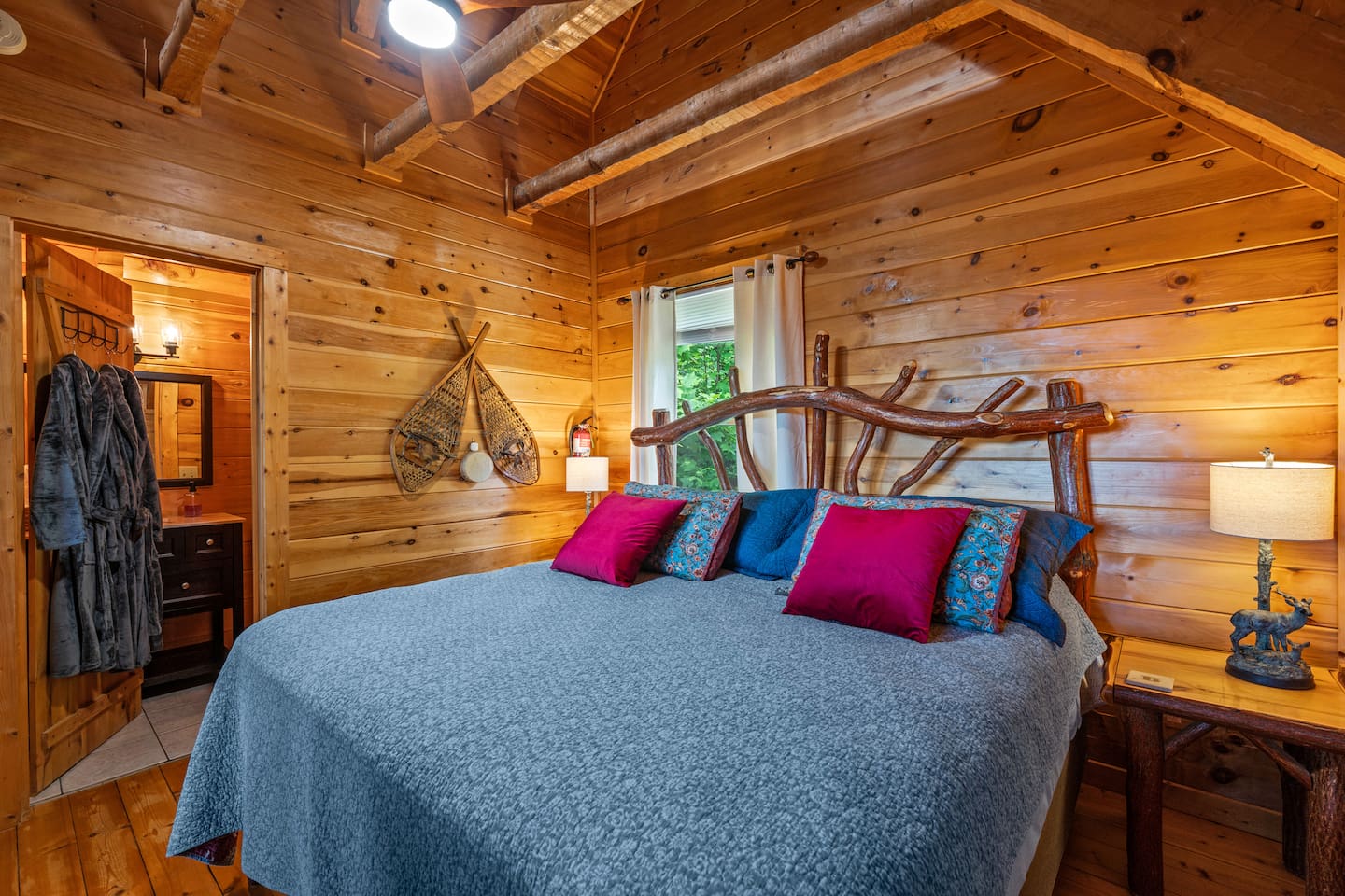 Cabin bedroom with a comfy King bed, colorful pillows, wooden walls, and a mounted TV. Red flowers on the dresser add a pop of color.
