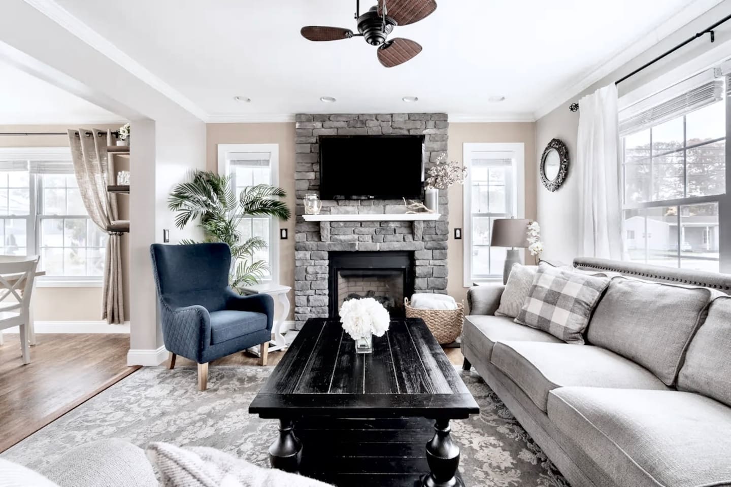 Relax in this inviting living space featuring a floor-to-ceiling stone fireplace, a large flat-screen TV, and a cozy sectional. Large windows and a ceiling fan create a bright, comfortable atmosphere for your stay.