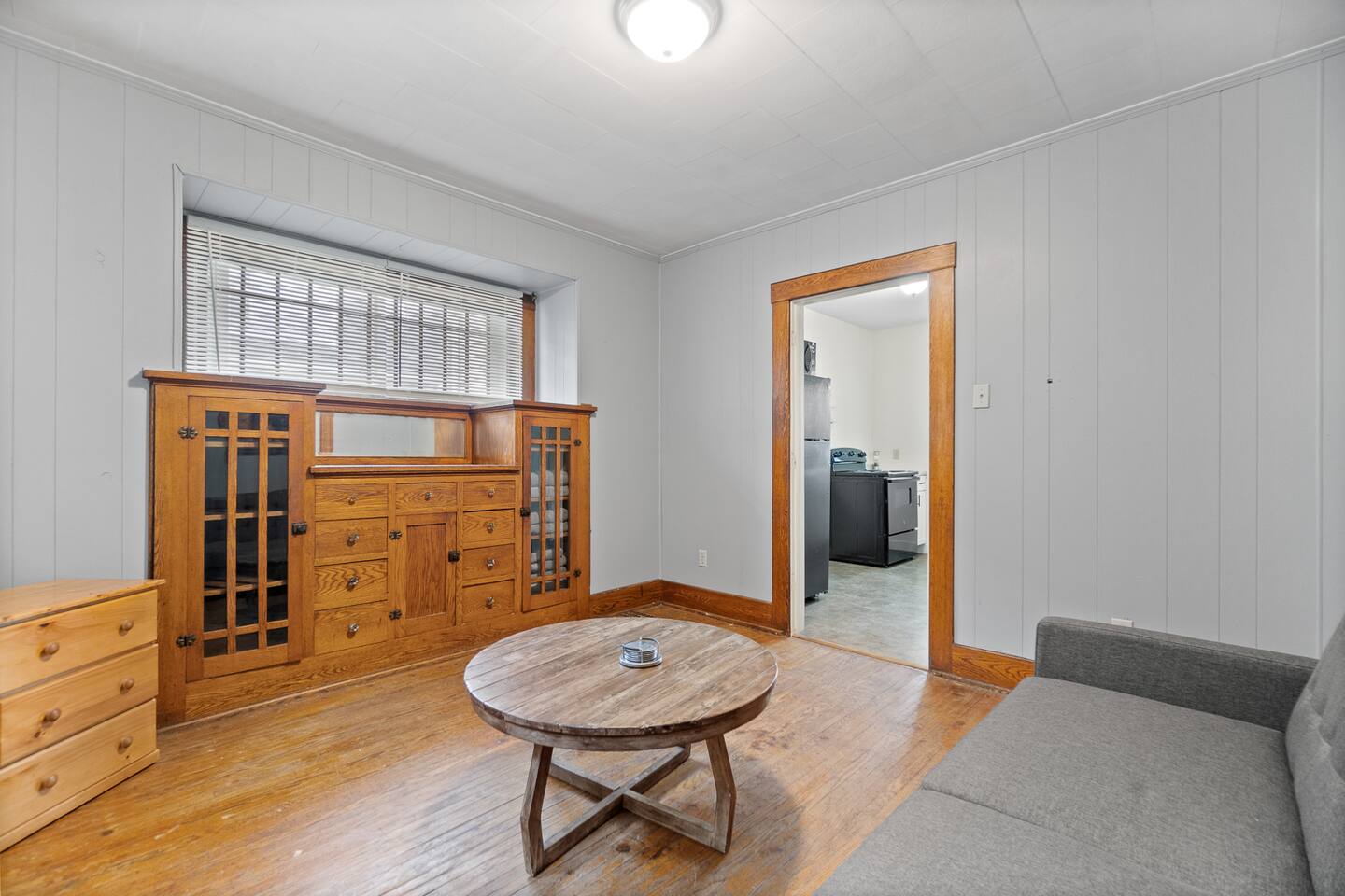 Cozy living room featuring a comfortable bed, stylish decorations, large windows, a tiny table in the middle, sleek wood flooring, a drawer, a cabinet, and a door leading to another room—creating a warm, inviting, and functional living space