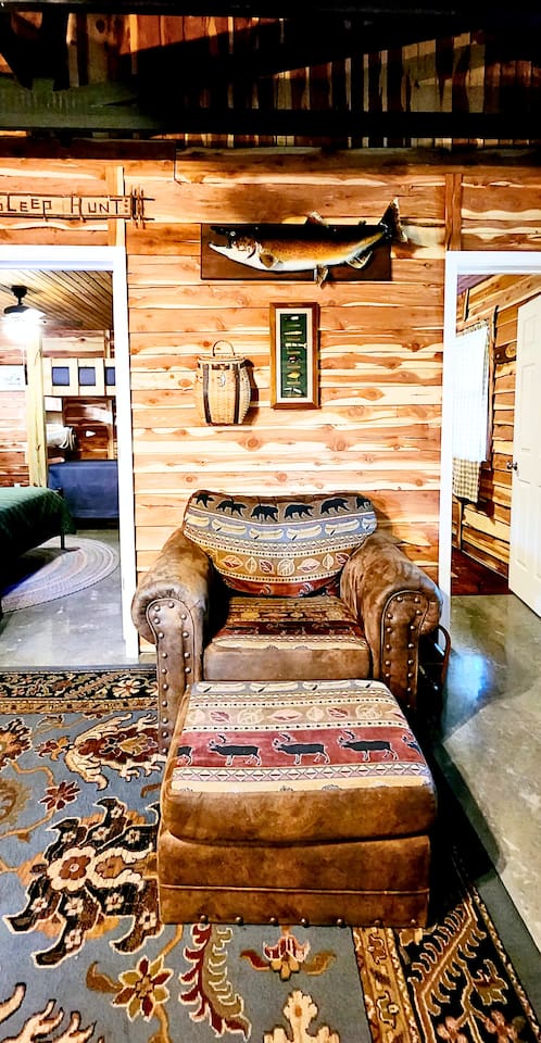 This oversized leather chair and footrest make the perfect reading or napping corner, accented with rustic wildlife patterns, soft rugs, and warm wood walls. Mounted fish decor and vintage touches bring classic lodge charm to this cozy spot.