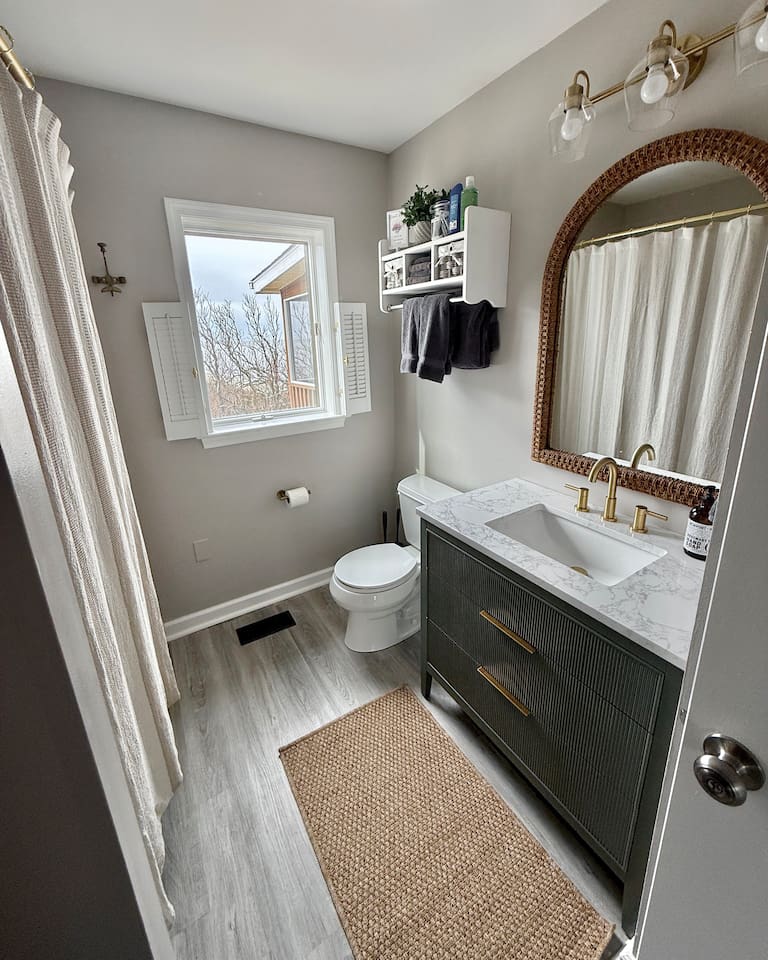 This clean, stylish bathroom features a modern dark vanity with gold hardware and marble countertops, paired with a woven rug and natural light from shuttered windows to create a serene, coastal atmosphere.