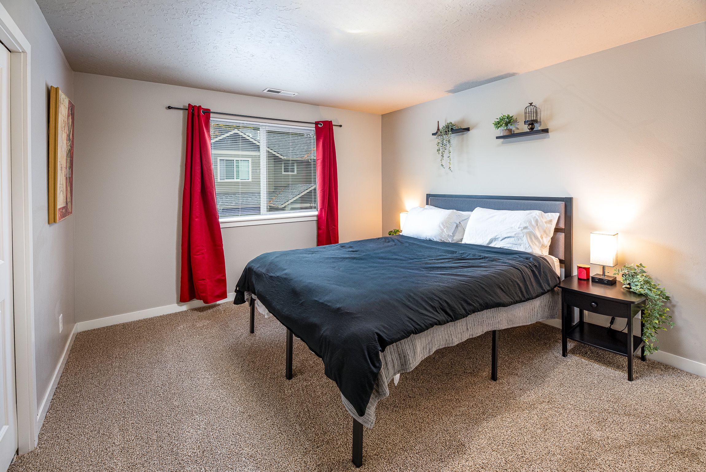 A cozy bedroom features a large bed with a black comforter and white pillows, flanked by two modern lamps on nightstands, red curtains framing a window that overlooks neighboring houses, and decorative shelves with plants on the wall.