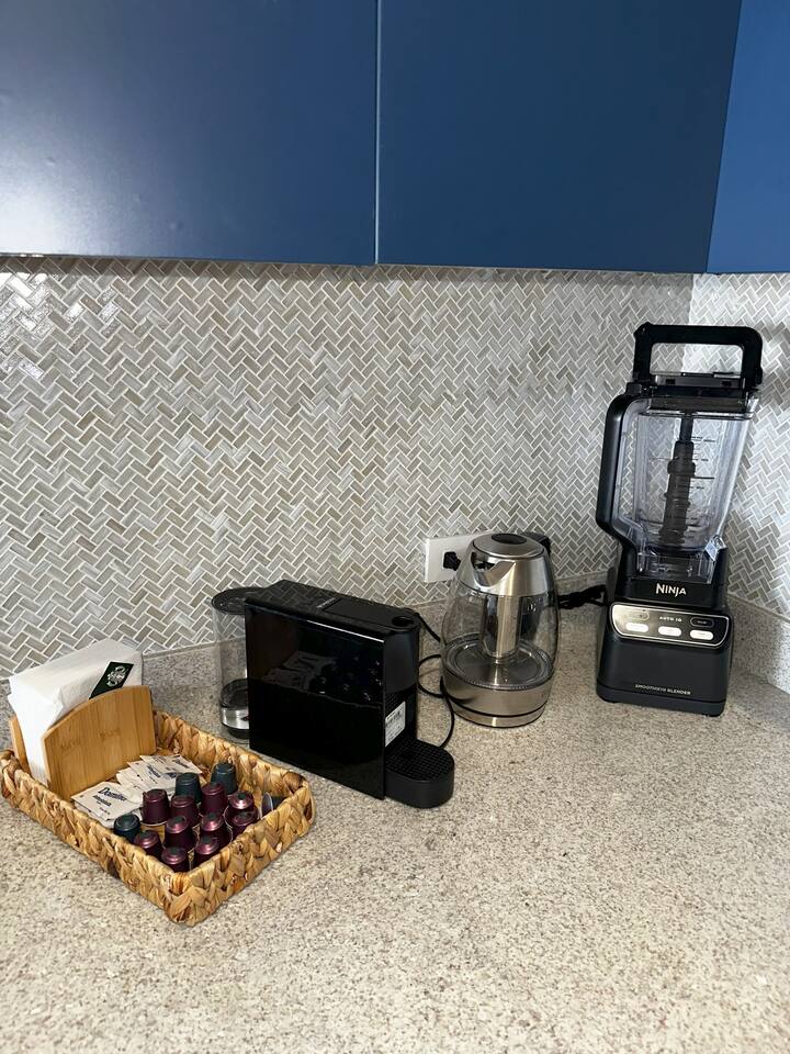 Elevate your mornings in this modern Cabo kitchen with a Nespresso Mini espresso machine, Sur La Table digital kettle with infuser, Ninja blender/smoothie maker, plus coffee pods, sugar, coffee, creamer, and tea—everything you need for your perfect drink.