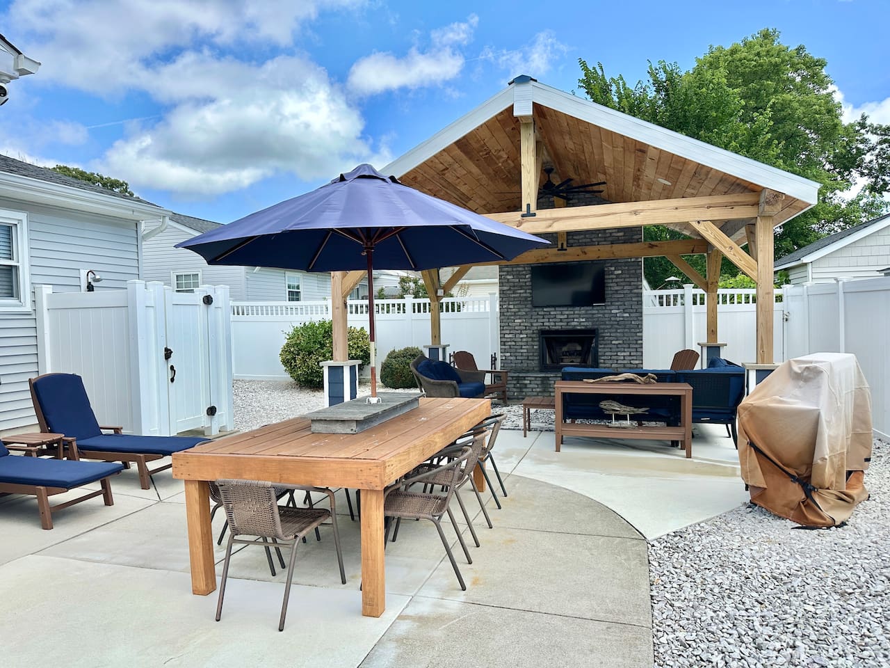 This backyard oasis features a massive wooden pavilion with a stone fireplace and wall-mounted TV. The space is fully equipped for relaxation and dining, including lounge chairs, a dining table with a blue umbrella, and a professional grill.