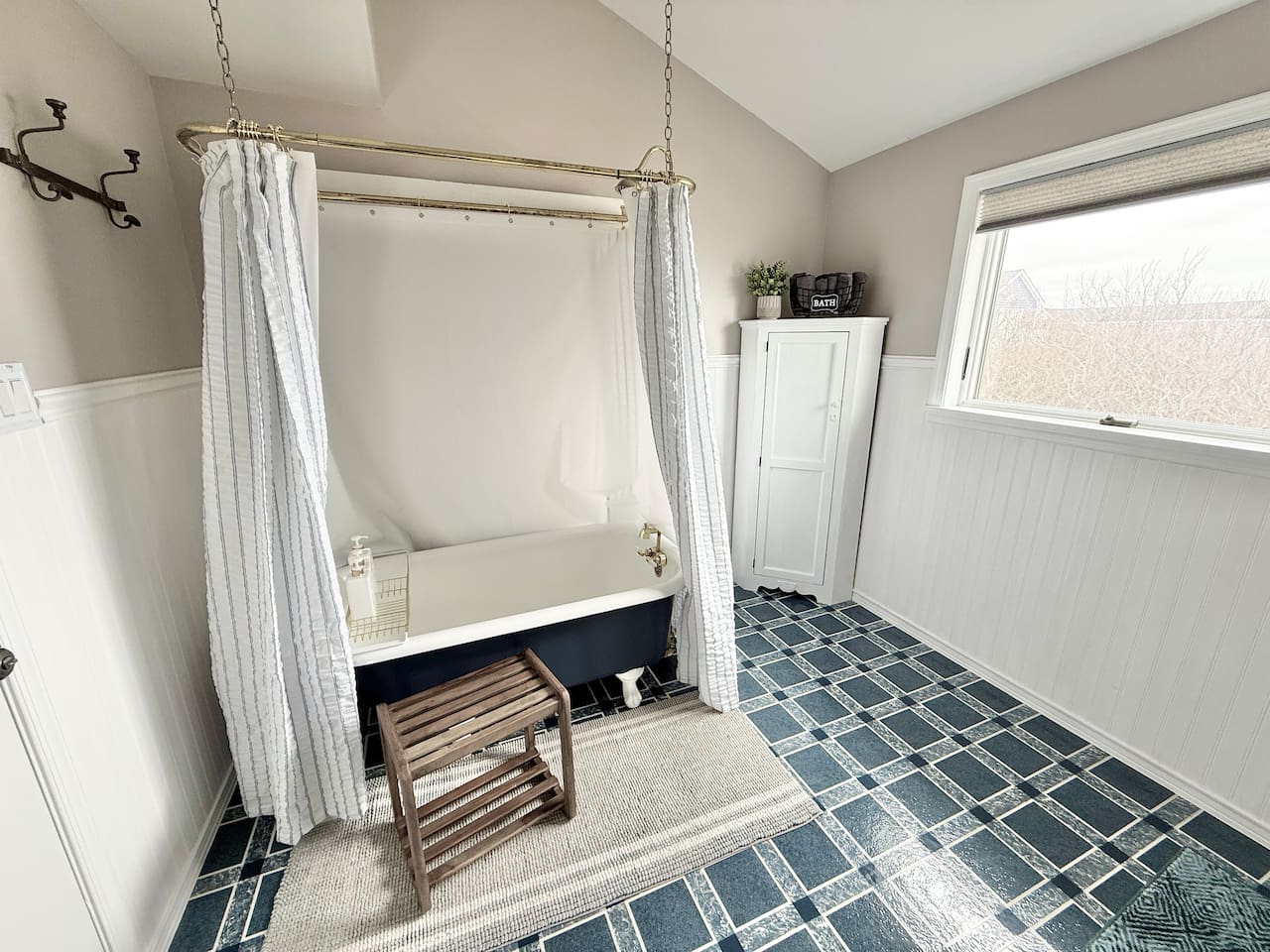 This spacious a coastal-style bathroom is highlighted by a clawfoot tub with a striped shower curtain, a wooden stool, and blue and white checkered tile flooring.