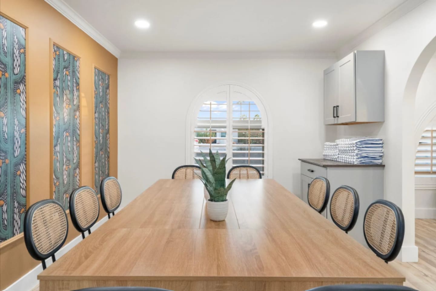 Gather the whole group around this oversized table, built for long dinners and even longer conversations. Woven-back chairs and vibrant wall art add warmth, while the sunlit archway window keeps the space feeling open all day.
