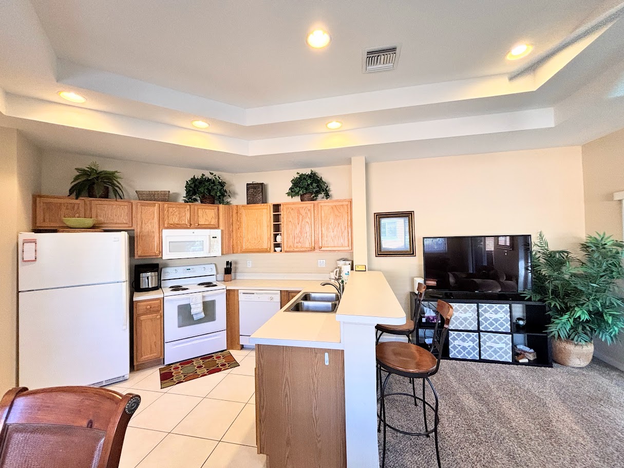 Open-concept kitchen and living area featuring full-size appliances, breakfast bar seating, large TV, and high tray ceilings for a bright, spacious feel.