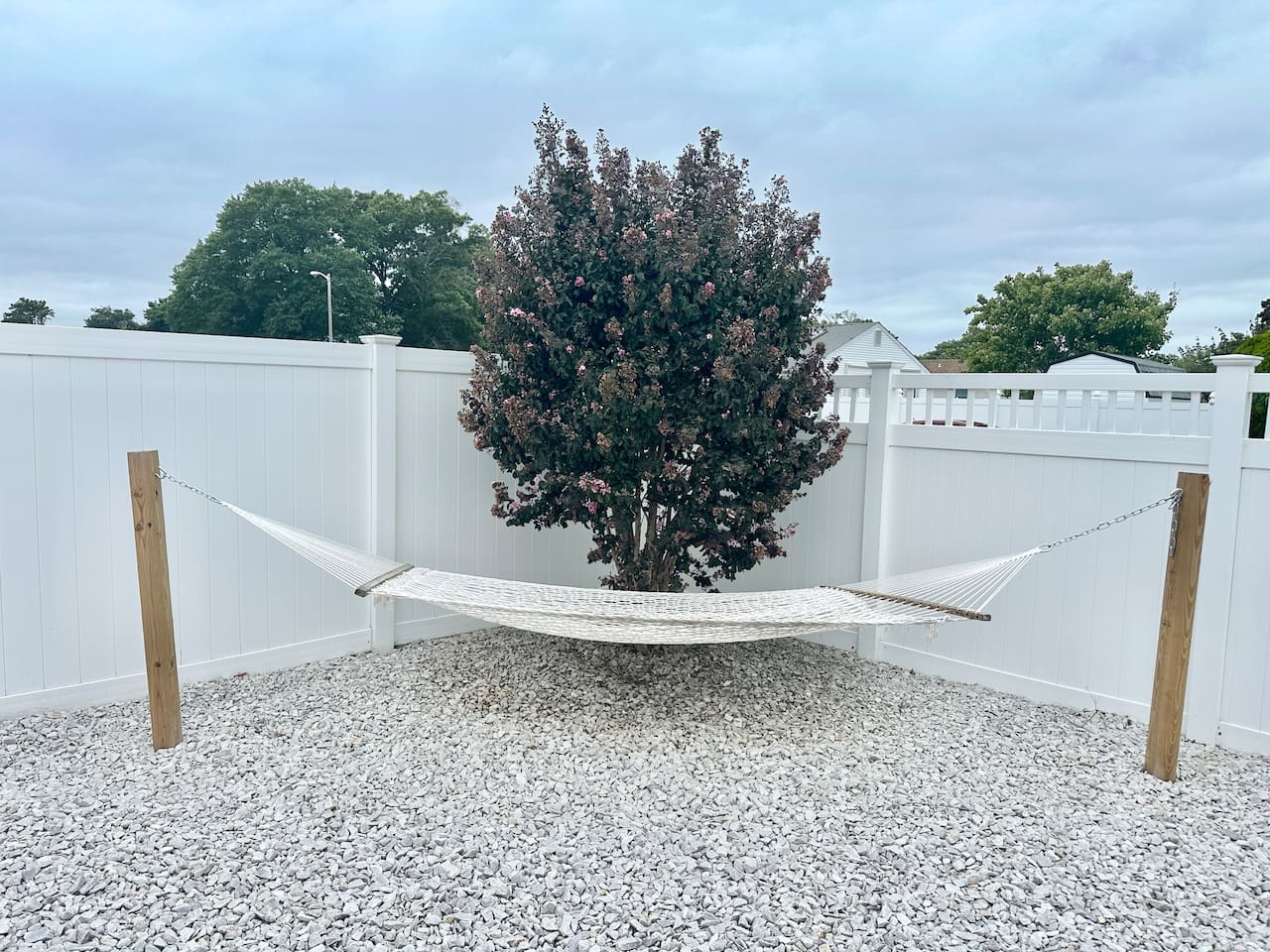 Escape to this peaceful corner of the private, fenced backyard, featuring a comfortable white rope hammock perfect for an afternoon nap. The hammock is set over a clean gravel base, offering a quiet spot to relax in the shade of a lush tree.