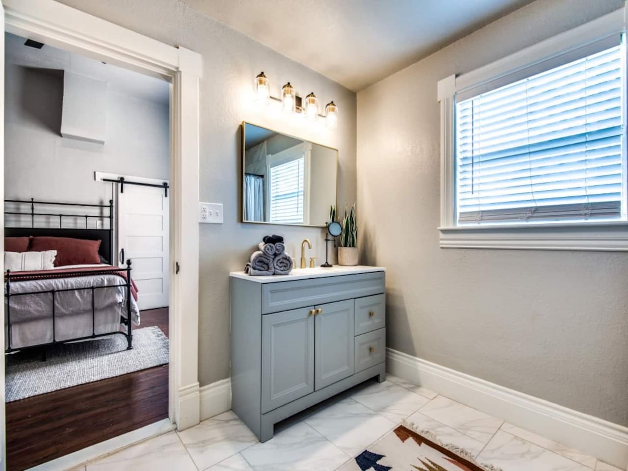 Bright vanity area with sage green double vanity, warm mirror lighting, wood-look tile floors, and a natural light window. Shared between Bedrooms 2 and 3 in this 3BR/2.5BA modern farmhouse Airbnb in downtown Aubrey TX.