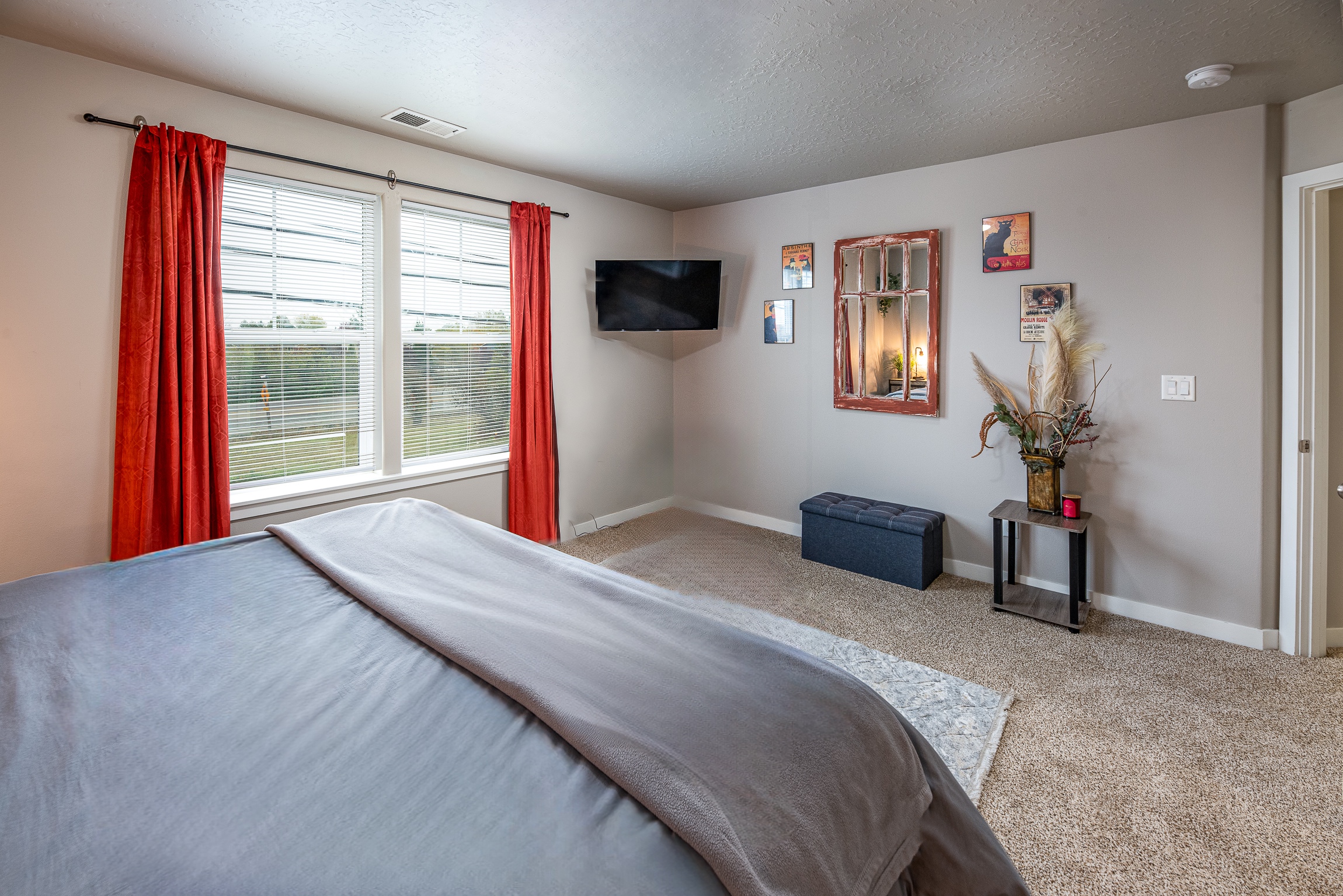 A cozy bedroom featuring a bed with gray bedding, bright orange curtains framing two windows, a mounted flat-screen TV, a decorative mirror on the wall, and a small seating area with a dark blue storage bench and a potted plant on a side table.