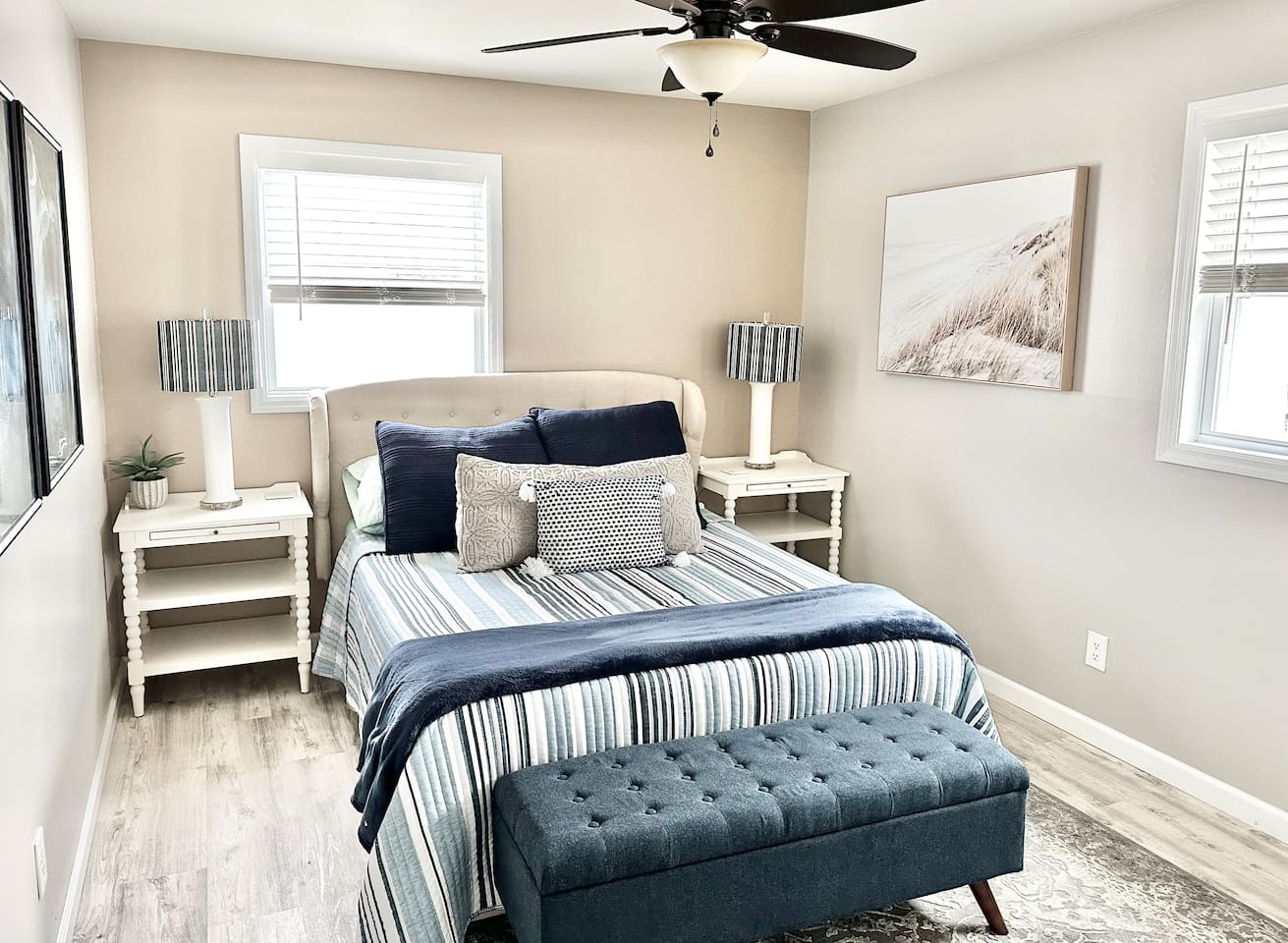 Relax in this stylish guest bedroom featuring a plush bed with nautical striped bedding and a tufted navy bench. With twin nightstands, coastal artwork, and plenty of natural light, it’s a bright and cozy retreat for your stay.