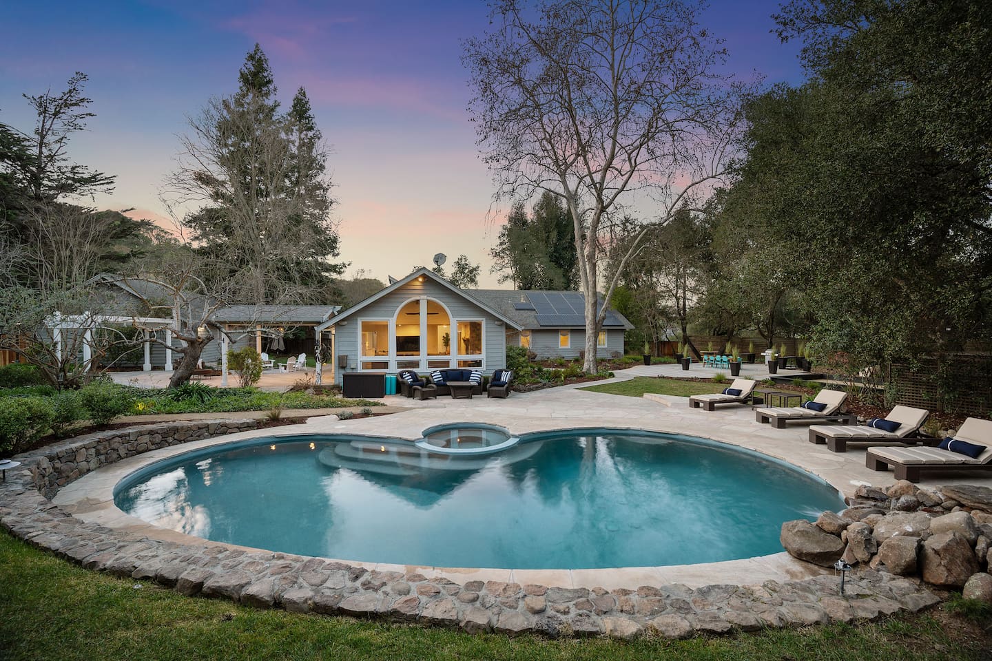 To the left, a pool table and seating area outside the kitchen and living room doors. In front, a pool, spa, rock waterfall, pool loungers, and couch + accent chair relaxation space. To the right, the dining pavilion and bocce court.