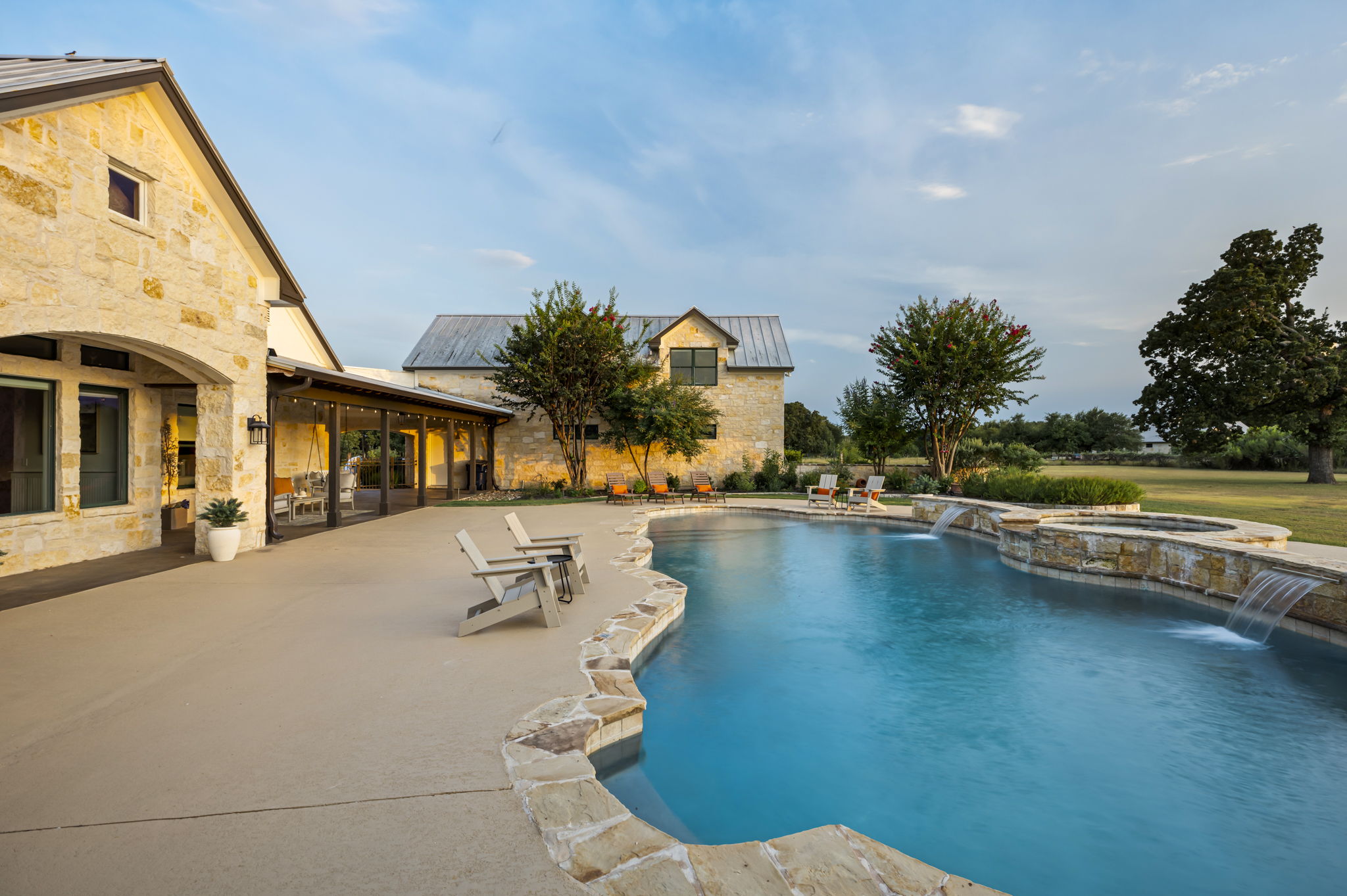 A luxurious Hill Country home featuring a sprawling outdoor living area with a resort-style pool, hot tub, and ample lounging space, set against a clear blue sky.