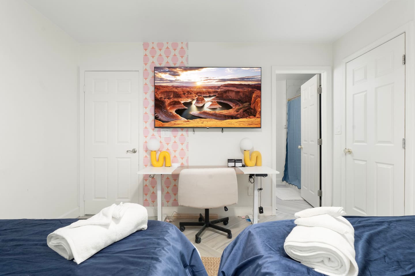Guest bedroom with dual beds, pink wallpaper, desk, and desert landscape art—comfortable space for couples or shared stays.