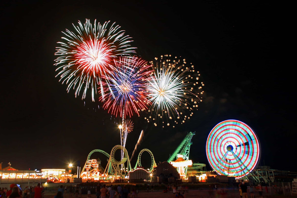 Nighttime fireworks over the boardwalk, with rides and lights illuminating the shoreline. A festive coastal scene where evening events, attractions, and ocean views come together for memorable nights by the beach.