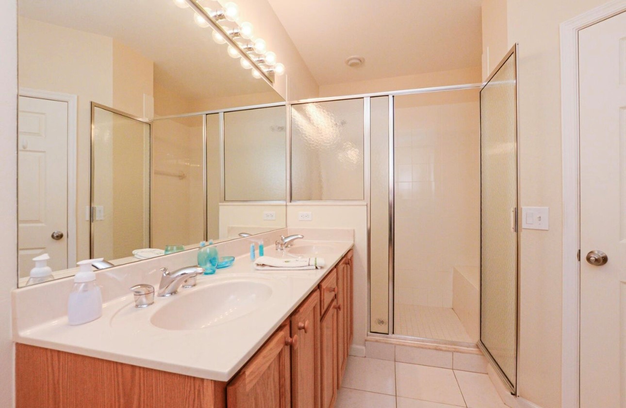Bright bathroom featuring a large vanity, full mirror, tile flooring, and a walk-in shower with glass doors.