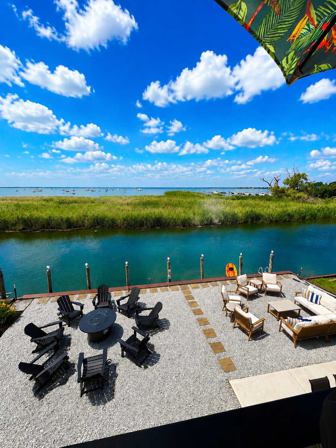 Wide waterfront patio with two seating areas Adirondack chairs around a fire pit and a lounge set overlooking calm canal water.