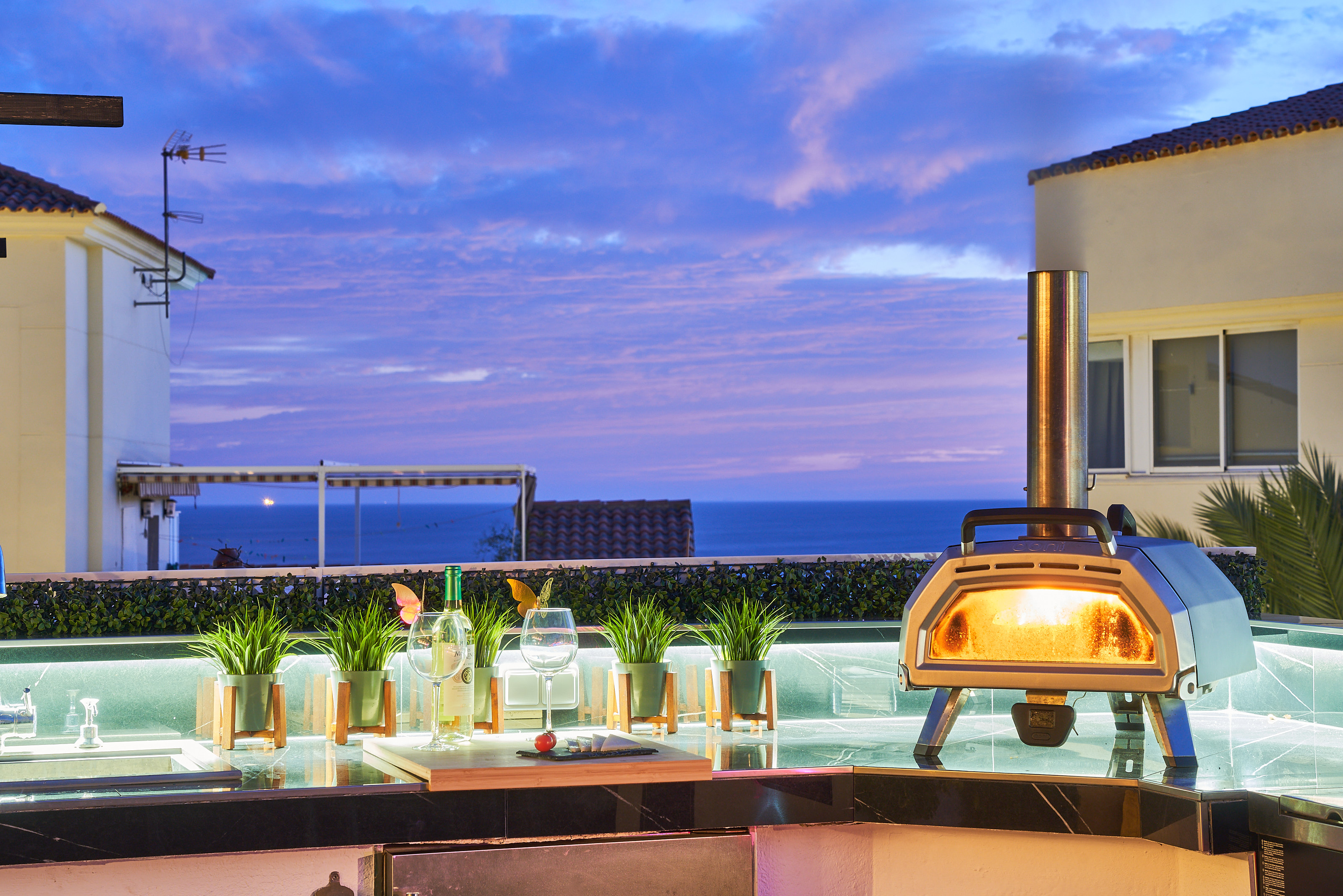 Fire up the built-in barbecue or the wood-fired pizza oven and enjoy cooking with a view. The fully equipped outdoor kitchen features a sink, fridge, and prep space with subtle lighting for evening meals. Dine under the open sky at the outdoor table, surro