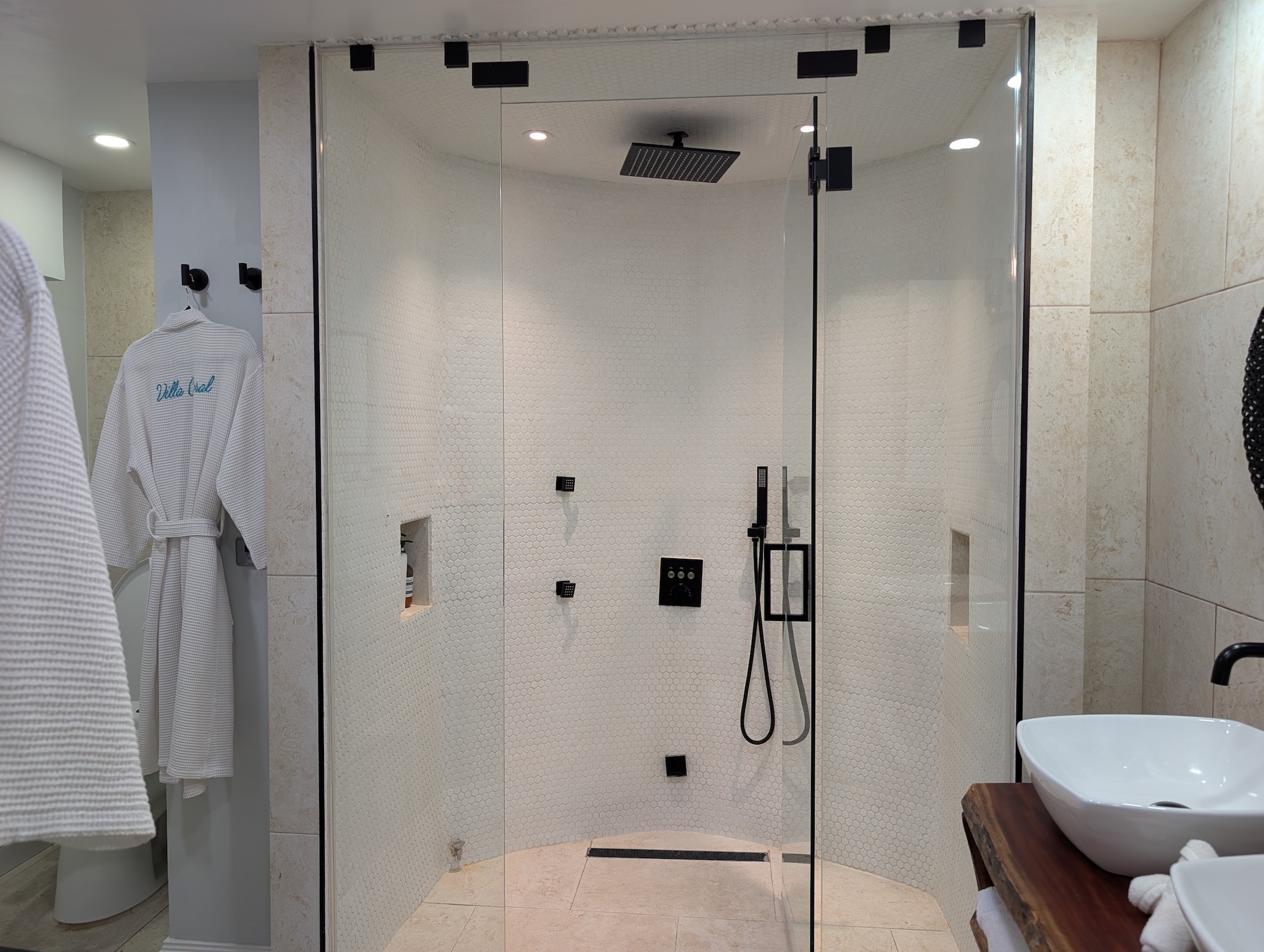 Glass-enclosed primary steam shower at Villa Coral featuring a 12-inch matte black rainfall showerhead, body jets, a handheld sprayer, white penny tile, and custom embroidered luxury robes in Dorado, Puerto Rico

