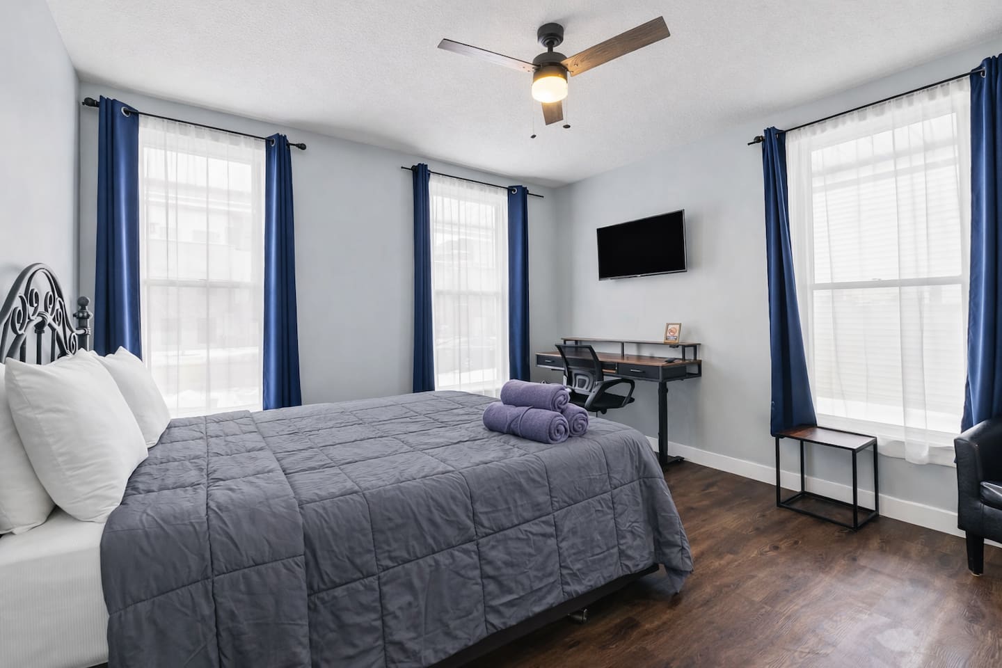 Bright bedroom view showcasing large windows, blackout curtains, ceiling fan, and a dedicated work desk.