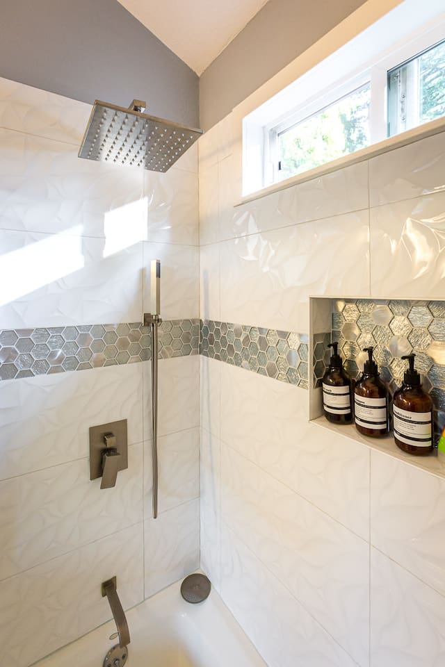 - Sleek shower with rainfall head, handheld sprayer & contemporary hardware finishes
- White textured tiles accented with metallic hexagonal band for a designer look
- Built in recessed shelf keeps bath products organized & within easy reach for use