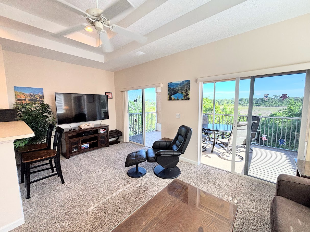 Bright living area with large TV, cozy seating, and sliding glass doors leading to the private screened balcony.