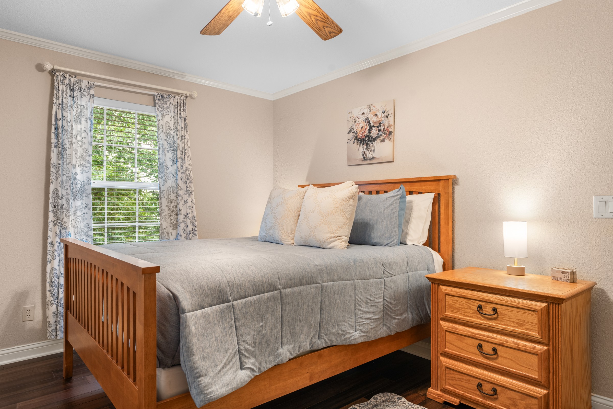 Main-level queen bedroom with oak bed and paisley curtains. Soft blue-gray bedding and a ceiling fan for easy sleep.