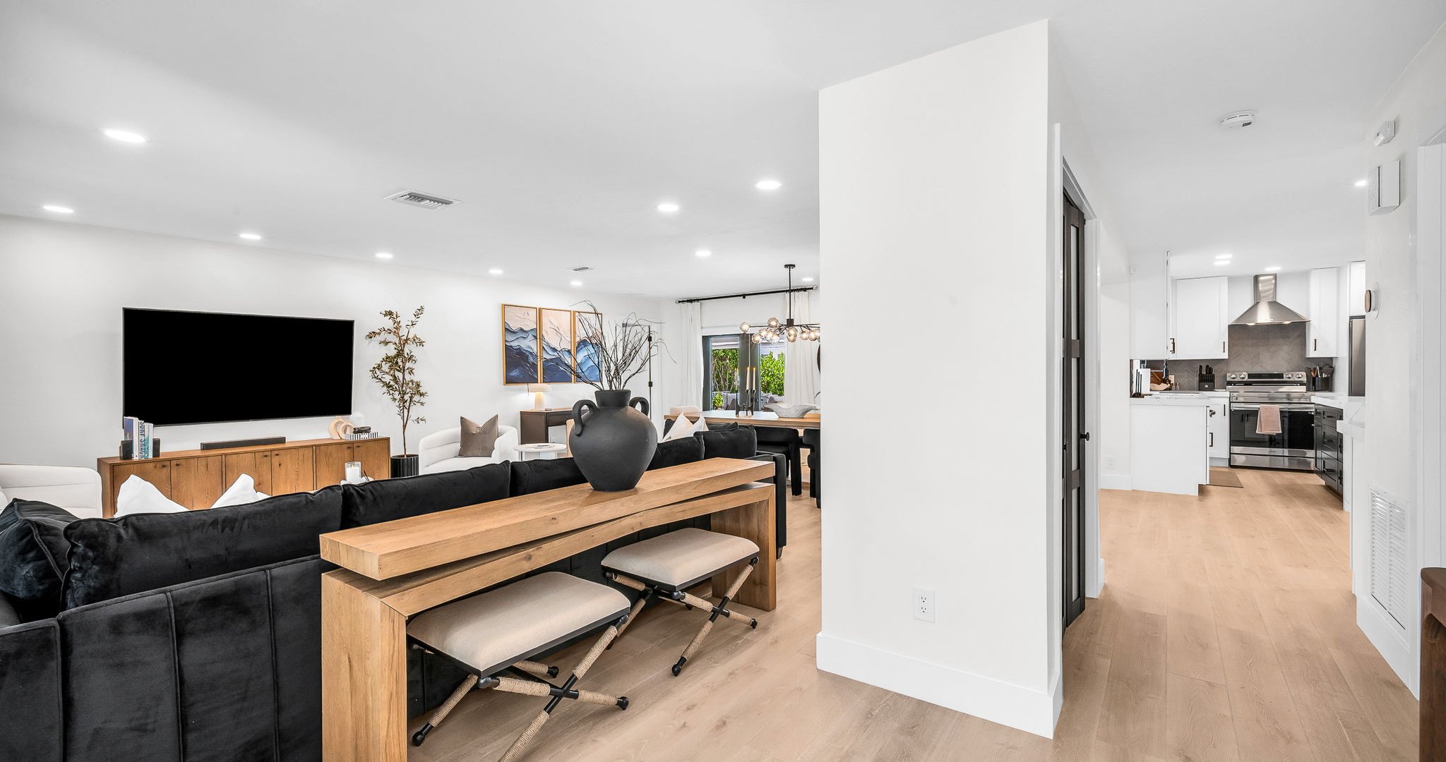 Elegant open-concept living room featuring a plush black velvet sectional, 75” smart TV, and designer mid-century modern furnishings. Floor-to-ceiling windows and French doors fill the space with natural light and connect seamlessly to the outdoor pool.