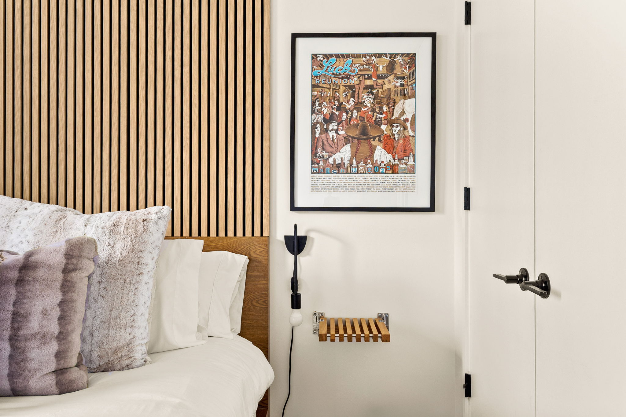 A cozy and modern bedroom corner featuring a unique wooden slat wall, a bed with plush pillows, a distinctive framed poster, and a sleek wall-mounted light fixture with a small floating shelf.