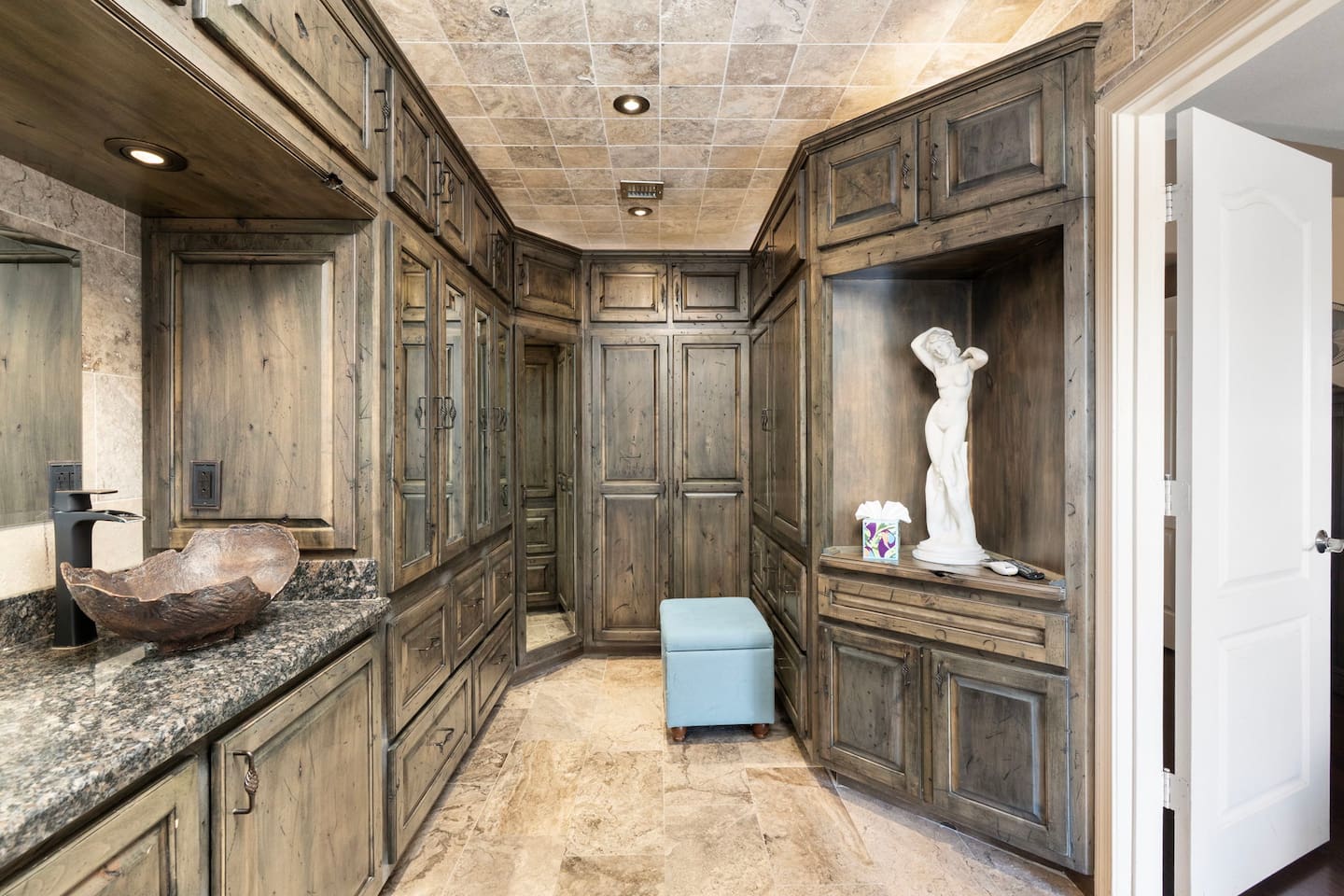 The walk-in closet is as beautiful as it is functional — wraparound distressed wood cabinetry, a vanity mirror, and a cushioned ottoman give you all the space and comfort to get ready in style.