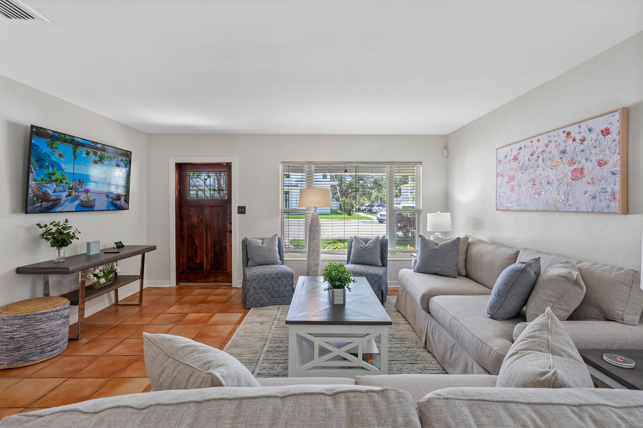 This sun-drenched second living room offers a breezy, relaxed vibe with its terracotta tile floors, light neutral furnishings, and large windows overlooking lush greenery.