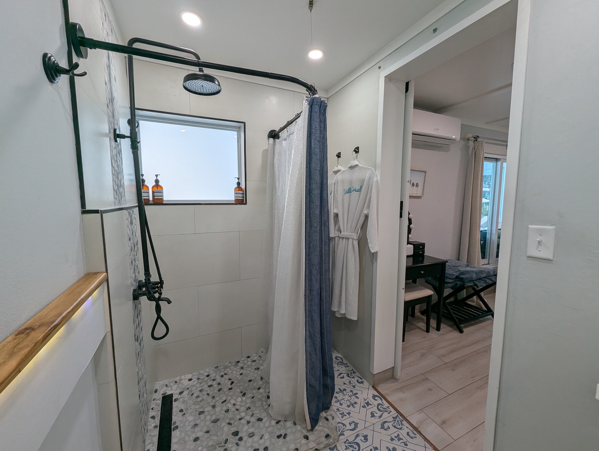 Walk-in guest shower at Villa Coral featuring a matte black rainfall showerhead, pebble tile flooring, custom tilework, and complimentary luxury robes in Dorado, Puerto Rico.