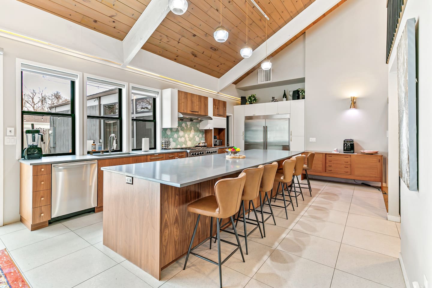 The kitchen is fully equipped with modern appliances, a Mid-Century Modern interior design, a large fridge, freezer, and ample storage. It boasts a spacious layout and a 16 ft island table, with convenient access to the outdoor area.
