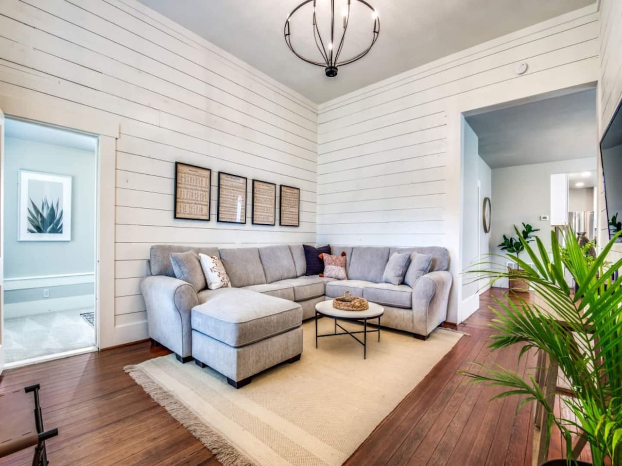 Corner view of the bright living room with full sectional, woven rug, hardwood floors, chandelier, and dual windows. Dedicated workspace available. Modern farmhouse Airbnb near Denton County wedding venues in downtown Aubrey TX.