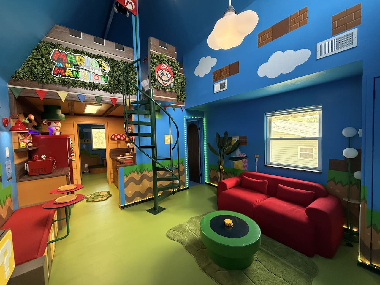 Explore the "castle" area with spiral staircase to the lofted Mario's Mini Mansion king bed! This vibrant blue living room features a squishy red sofa, a green coffee table, custom signage, and playful Mario details -- endless adventures for all.