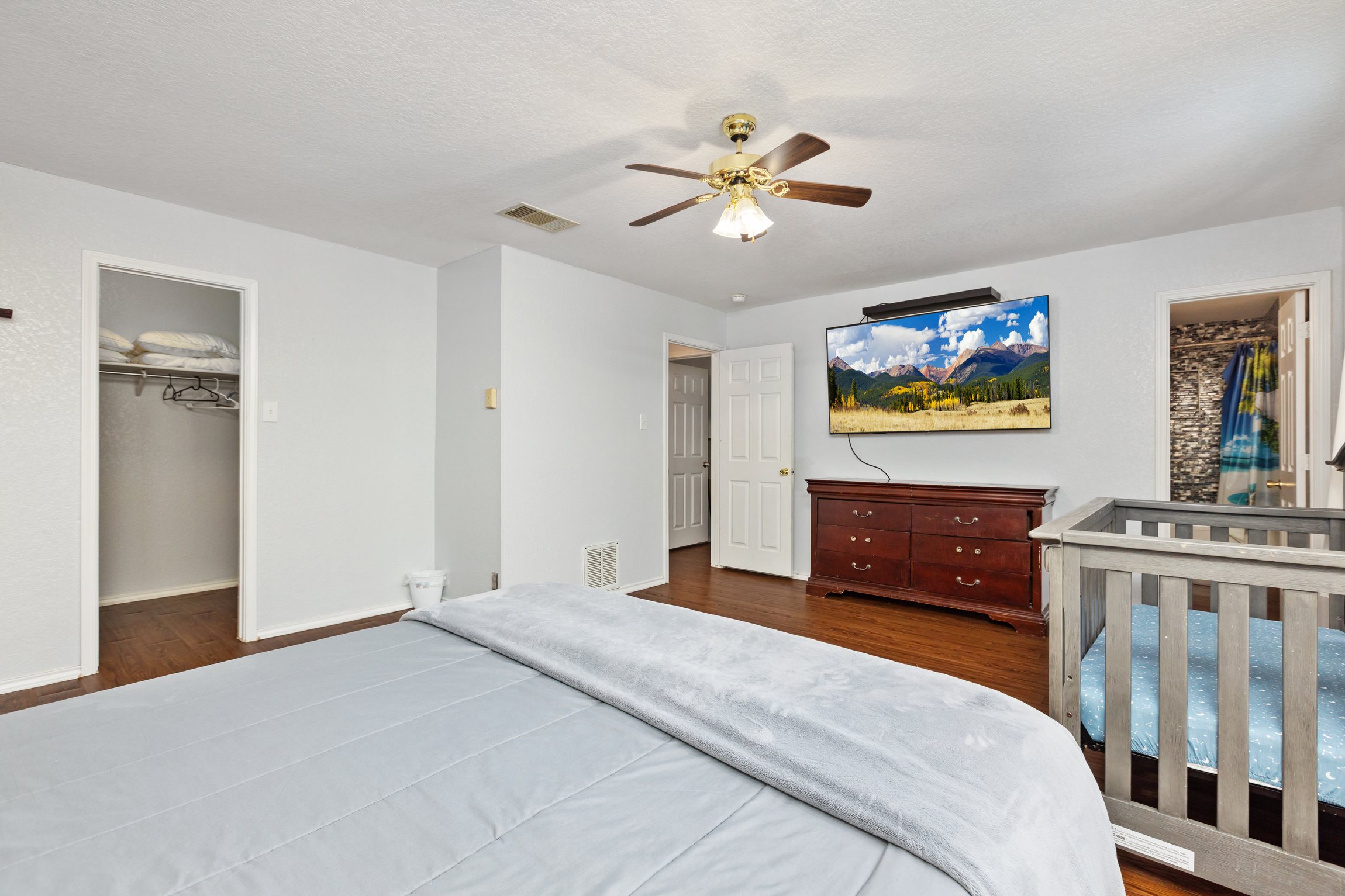 Bedroom with wall-mounted Smart TV, streaming, ceiling fan, clothing storage, and quality bedding. Room-darkening shades and extra pillows included. Baby crib conveniently located in the primary bedroom.