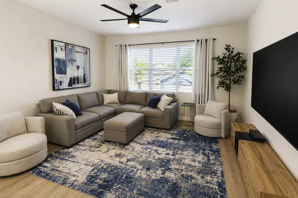 Spacious and inviting living room with a plush sectional and ottoman, perfect for lounging or gathering. Enjoy abundant natural light, a large TV for entertainment, and a cozy seating area designed for comfort and relaxation.