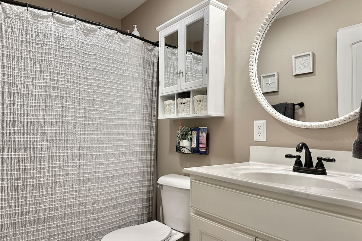 This bathroom features a white vanity with a dark faucet and a large, beaded-frame round mirror. It includes a white wall cabinet with glass doors, a standard toilet, and a shower area with a textured grey curtain.
