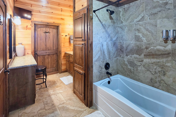 Private bathroom inside primary bedroom with a tub/shower combo, warm wood finishes, stone-look tile surround, 2 vanity areas, and plenty of cabin character. Hollywood lighted mirror available under counter.