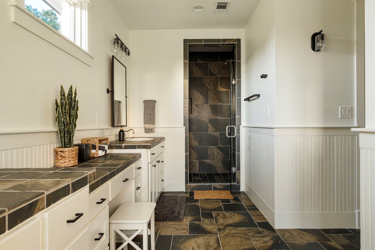 A walk-in shower with dark tiled walls and floor, creating a striking contrast with the bright white vanity area.