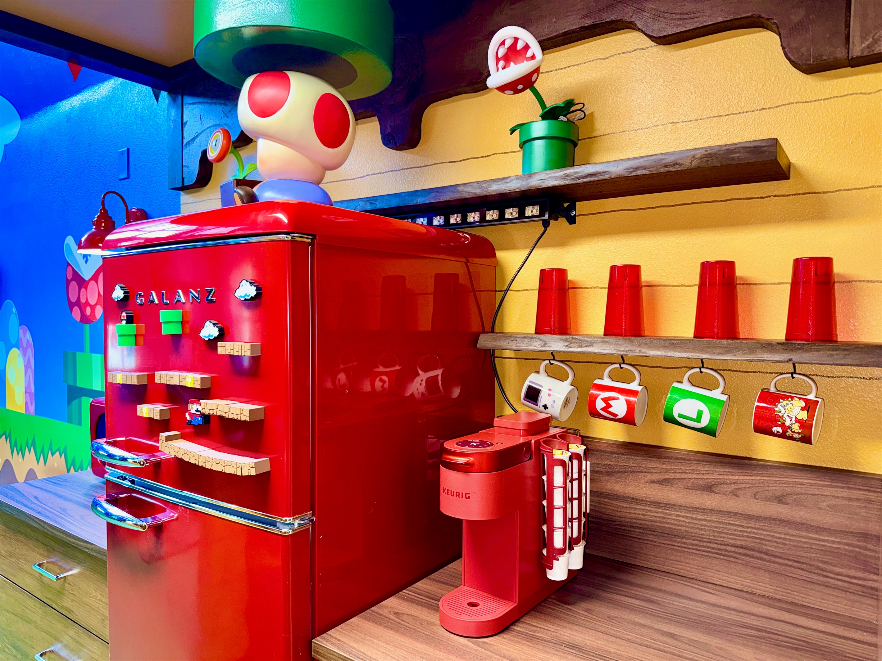 Whimsical kitchen at Mario's Mini Mansion, Clermont, FL! Bright red retro-style fridge, green flooring, yellow walls with Mario shelves, and Toad-inspired decor make post-theme park meal prep an adventure for kids and adults alike.