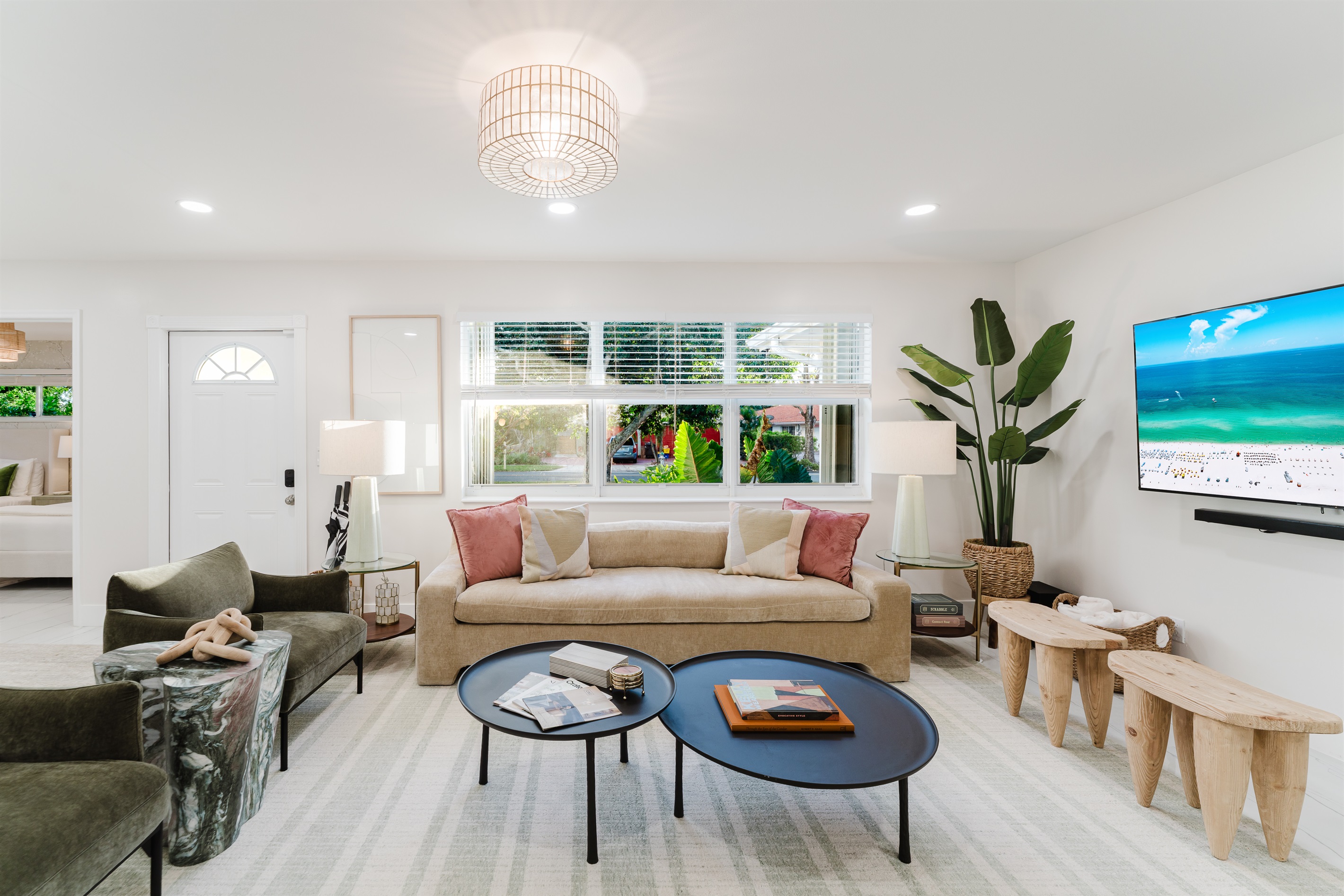 Light-filled living room with a 65" smart TV, sound bar, and subwoofer, featuring plush designer seating, neighborhood views, curated games, and reading materials—perfect for relaxing, entertaining, and family movie nights in West Palm Beach.