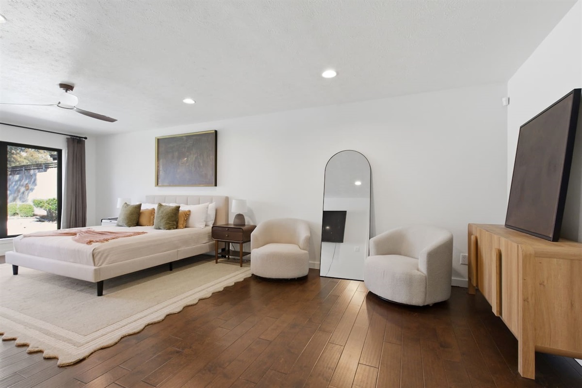 Huge primary bedroom featuring a king bed, 65” Smart TV, ceiling fan, cozy sitting area, full-length mirror, room-darkening shades, and generous dresser storage space, with direct access to the backyard for added comfort and convenience.