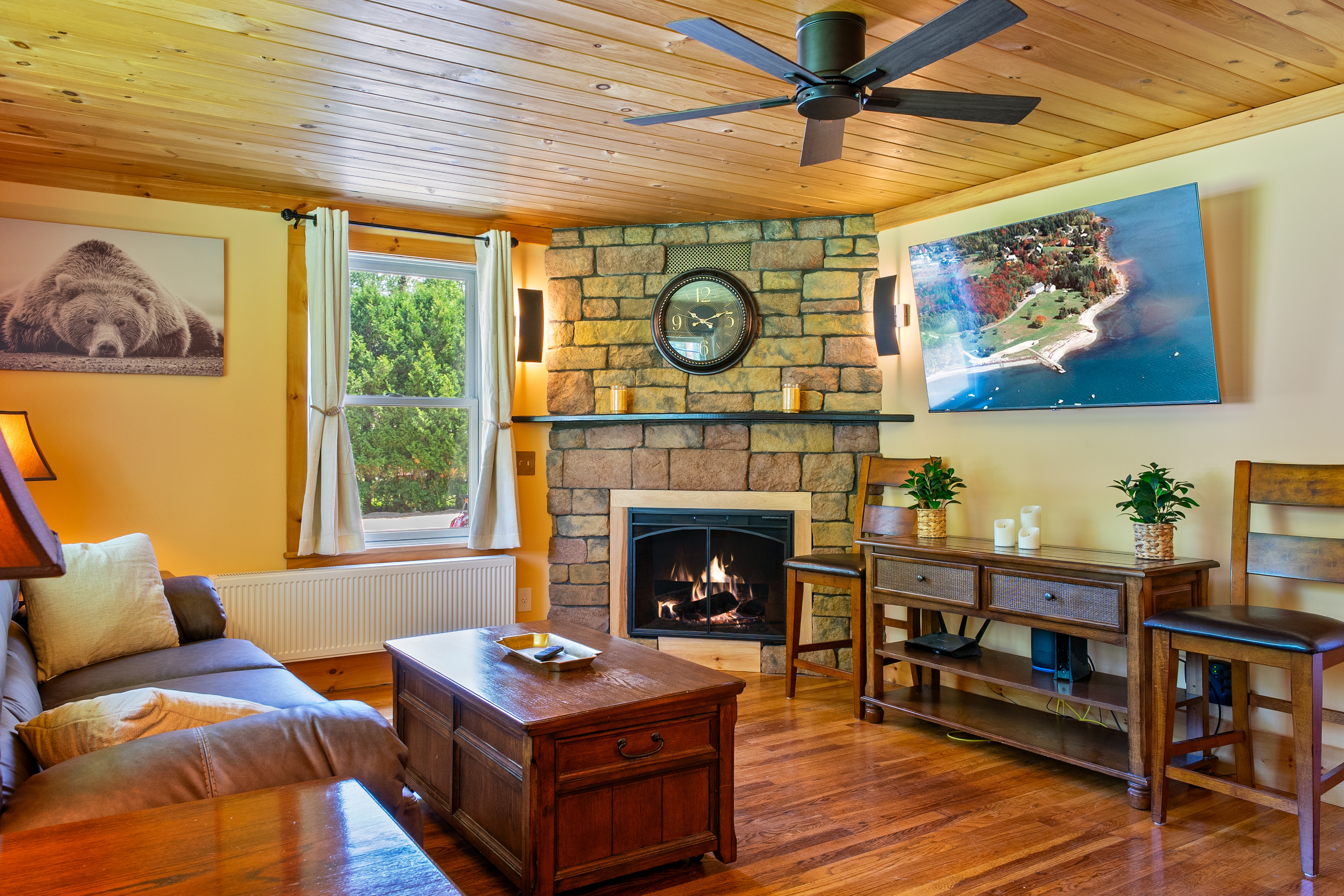 Gather in the main living room featuring a warm stone fireplace, natural pine ceilings, and nature wall art. Stream your favorite shows on the wall-mounted Roku smart TV while enjoying comfortable seating after a day exploring coastal Maine.