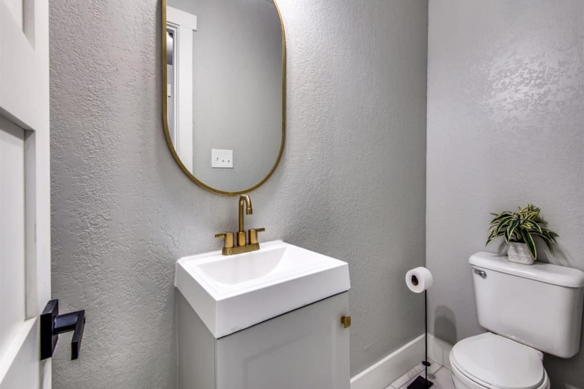 Stylish powder room off the kitchen with white pedestal sink, gold fixtures, and oval mirror. Added convenience for groups up to 7 in this 3BR/2.5BA modern farmhouse Airbnb in historic downtown Aubrey TX.