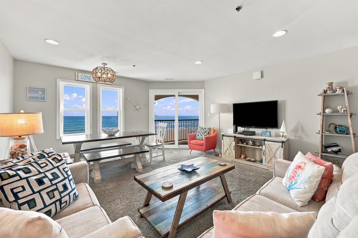 Inviting open-plan living room arrangement showcasing plush neutral sofas adorned with vibrant coastal accent pillows, a rustic wooden coffee table, a sleek media console, and a seaside dining area set against sweeping ocean views.