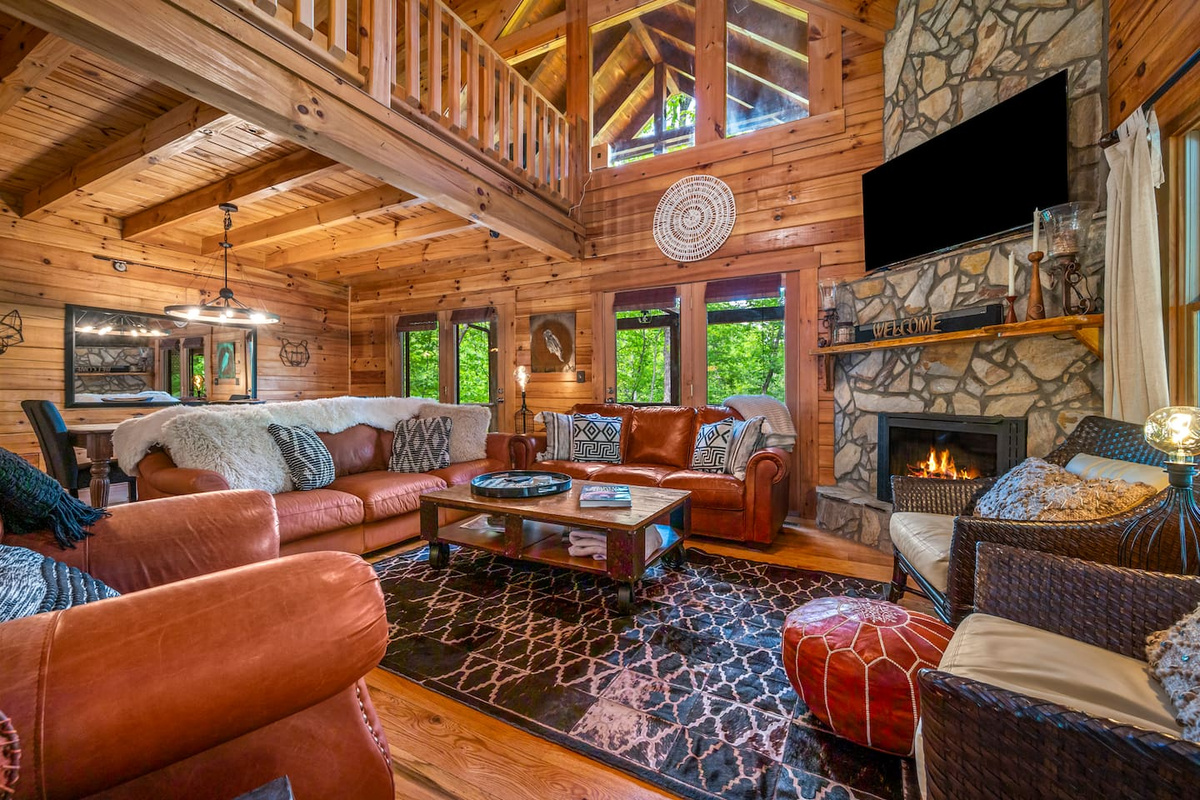 The heart of the cabin features a main living room with soaring, vaulted ceilings, warm wood interiors, plush seating, and a stunning stone fireplace. Large windows bring in the natural outdoor light, creating the perfect space to relax.