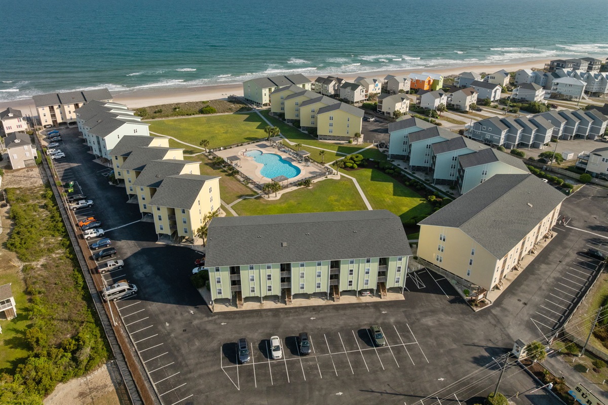 Elevated aerial capturing beachfront buildings, pool area, and lush green space