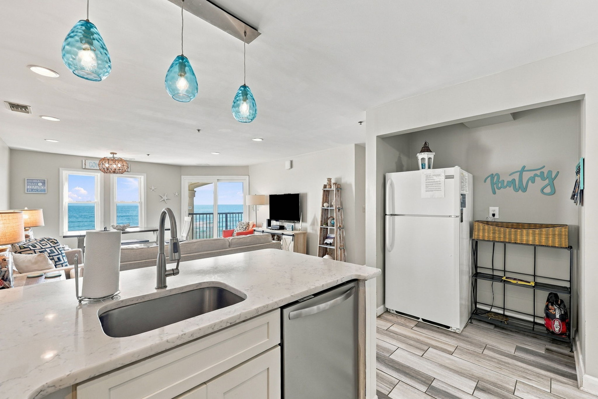 Panoramic perspective from behind the kitchen counter highlighting the open layout, with vibrant turquoise blown-glass pendant lights, an undermount stainless steel sink, a recessed refrigerator and pantry alcove, and floor-to-ceiling vistas of the turquoi